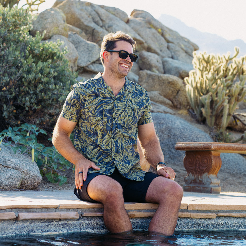 Man wearing Kantis Shirt with leaf pattern, seated by pool with legs in water, sunglasses, beach-inspired comfort apparel.