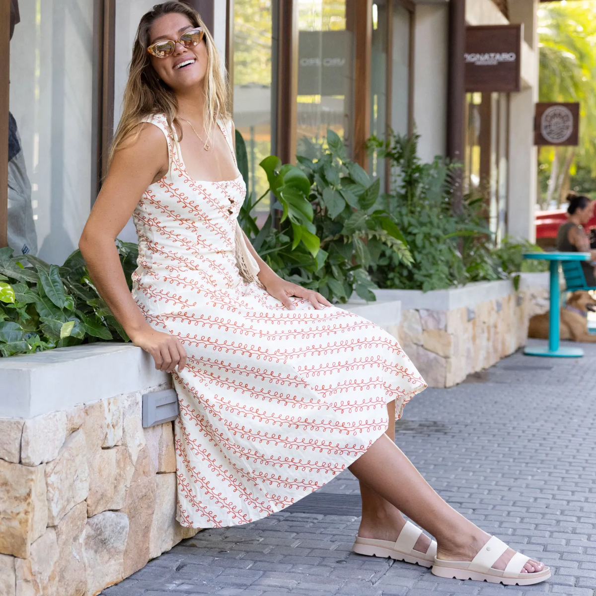 Woman wearing Vista Luxe Thea sandals facing right, seated outdoors with plants and stone wall background, beach-inspired comfort shoes.