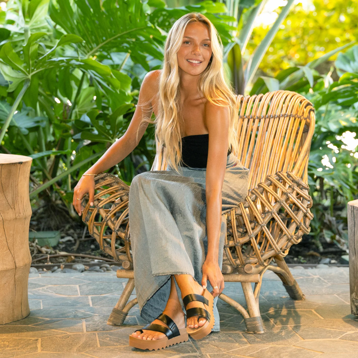 Woman seated on wicker chair wearing black Vista Hi Layla sandals facing forward, showcasing comfort and beach style