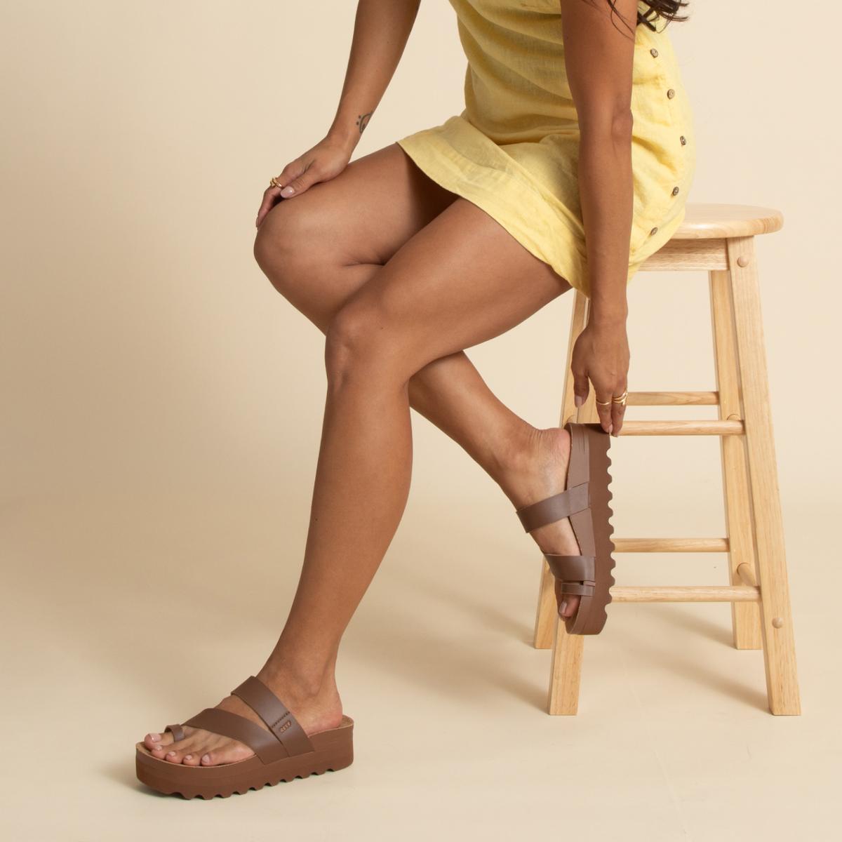 Woman wearing brown Vista Hi Layla sandals facing right, sitting on a wooden stool, showing footbed and arch support.