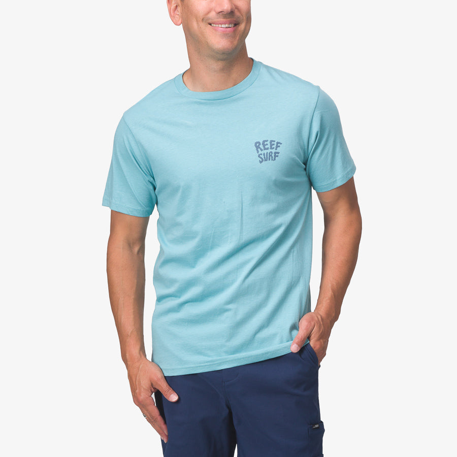 Man wearing light blue La Loca Tee with reef surf logo on chest, hands in pockets, smiling, front view.