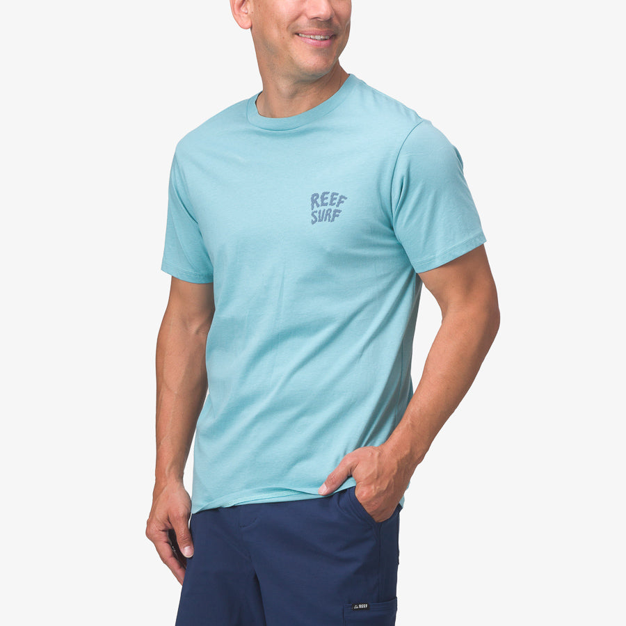 Man wearing light blue La Loca Tee with REEF SURF print, front view, styled for beach comfort and durability.