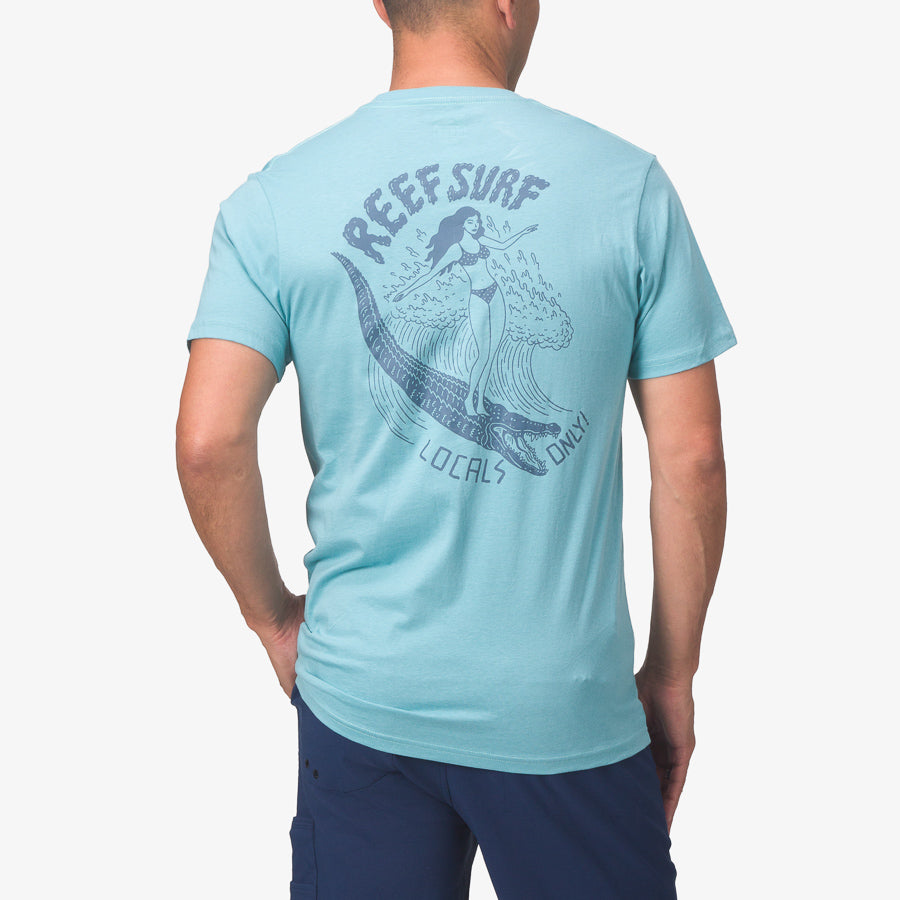 Man wearing light blue La Loca Tee with surf-themed graphic on back, standing with hands on hips, back view.