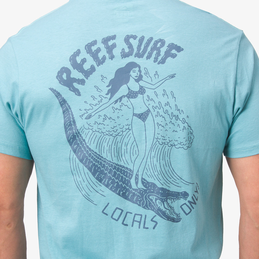 Back view of a man wearing La Loca Tee in blue with surf-inspired graphic of woman and crocodile on the back.