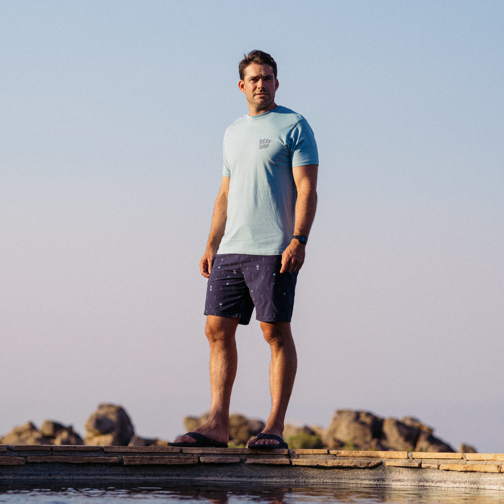 Man wearing La Loca Tee in light blue, standing outdoors in beach-inspired casual apparel with sandals facing forward.
