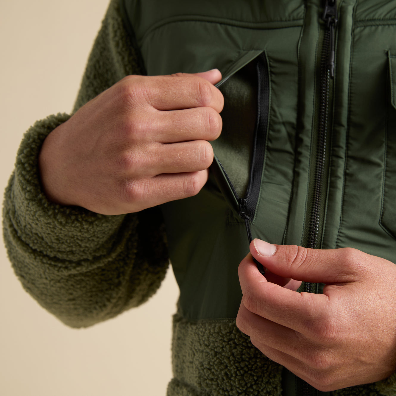 Close-up of hands unzipping chest pocket on olive green Cooke Sherpa Jacket showcasing durable fabric and comfort features.