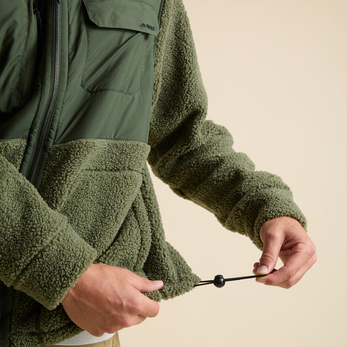 Close-up of hands adjusting the hem drawcord on the olive green Cooke Sherpa Jacket with textured fabric front panel.