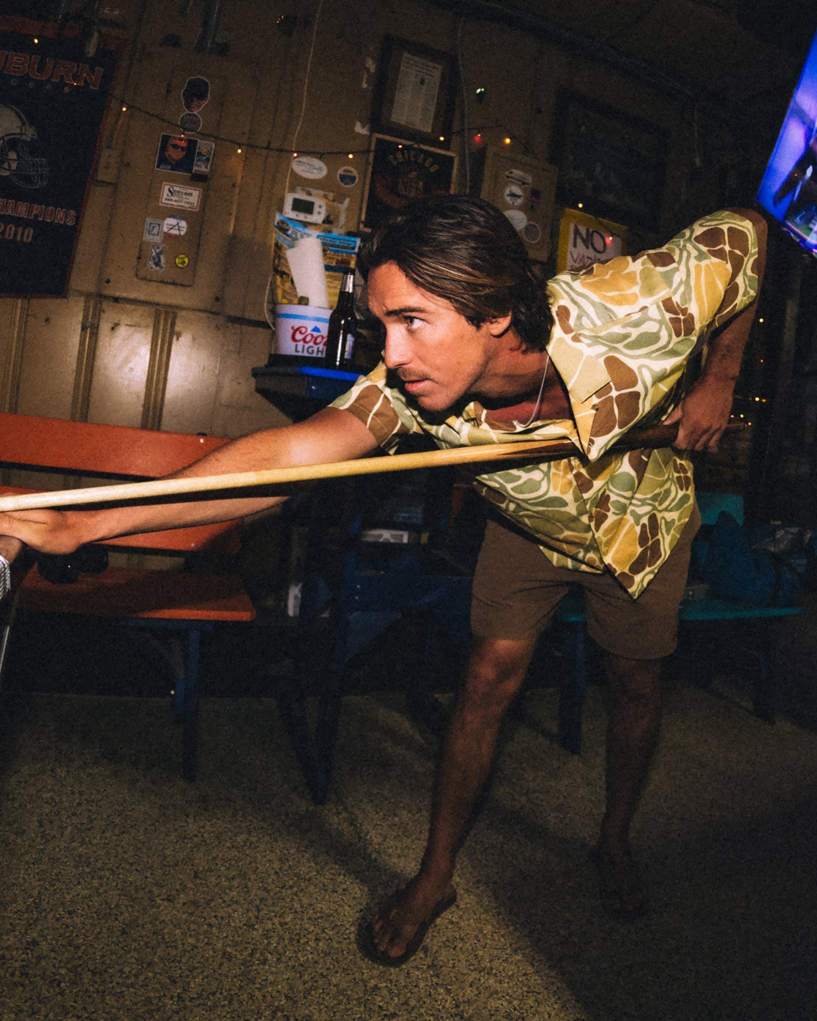 Man playing pool indoors wearing Men's Sullivan's Camp Shirt - Floral Camo with brown shorts and barefoot facing left