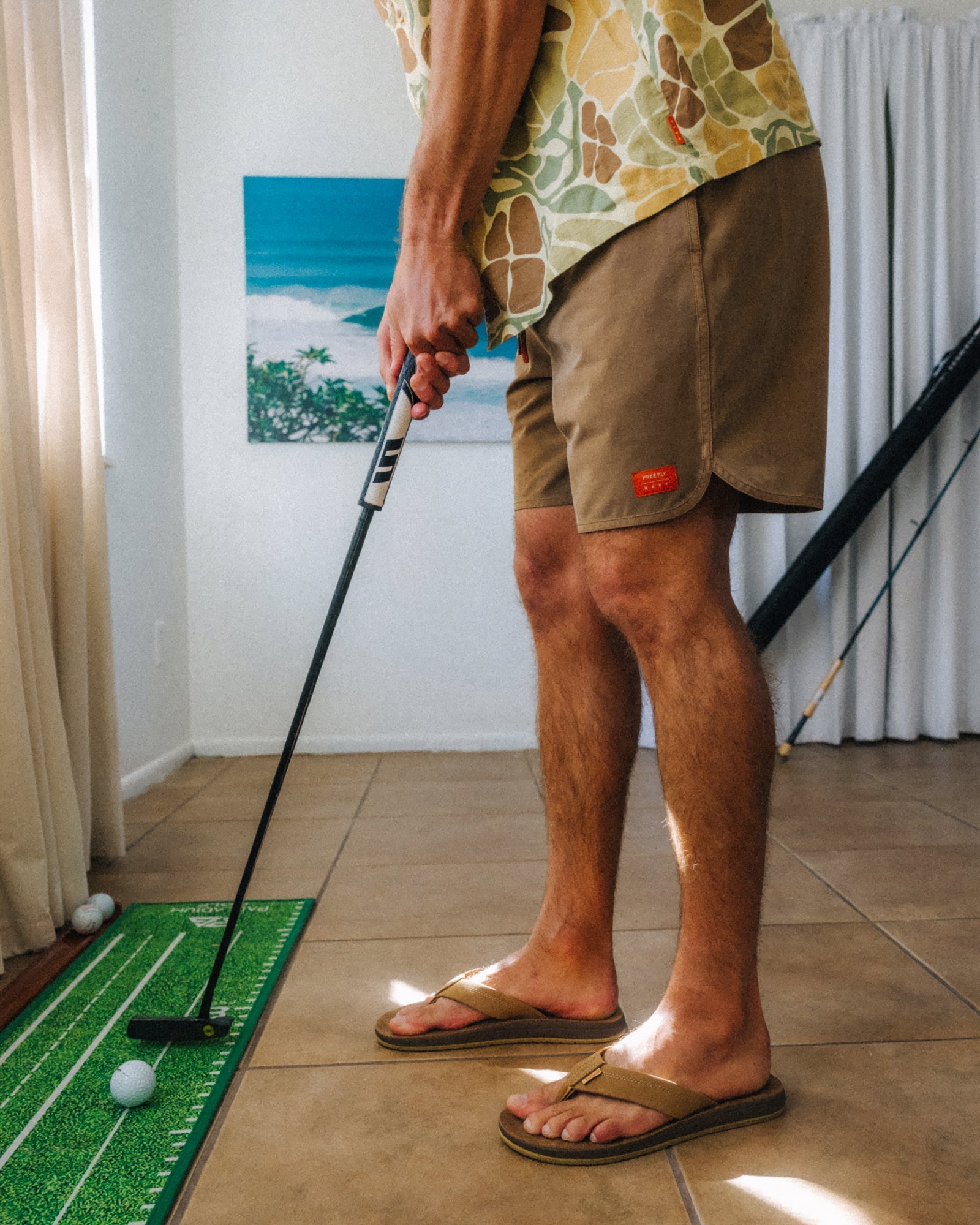Man wearing Men's Reverb Short Unlined – 7" - Coconut with tan sandals facing right indoors playing mini golf on green mat
