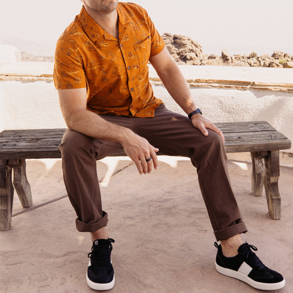 Man wearing Matthew Twill Pant seated on wooden bench, showing front view with rolled cuffs and black sneakers facing forward.