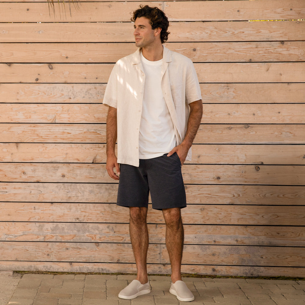Man wearing Neptune Slip-On shoes facing forward, paired with casual beach-inspired outfit and wooden background.