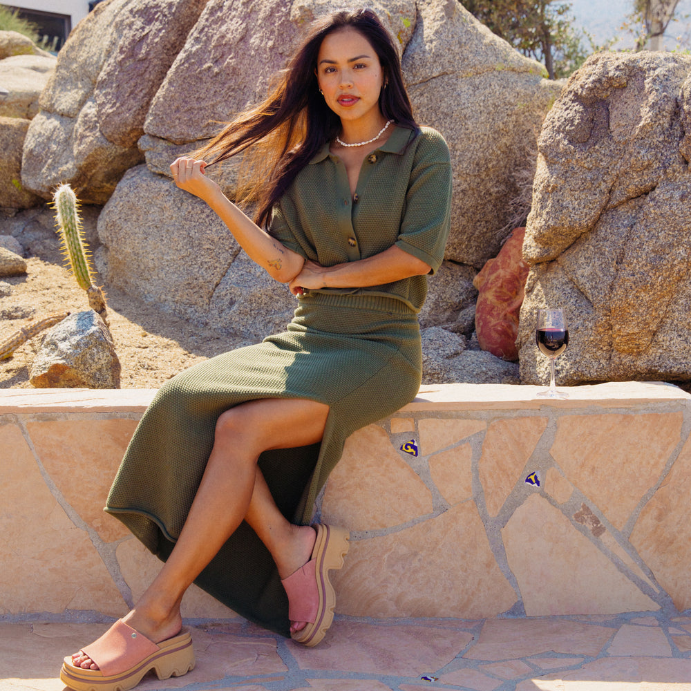 Woman seated outdoors wearing olive Up North Sweater Skirt with legs crossed and front of beige platform sandals visible