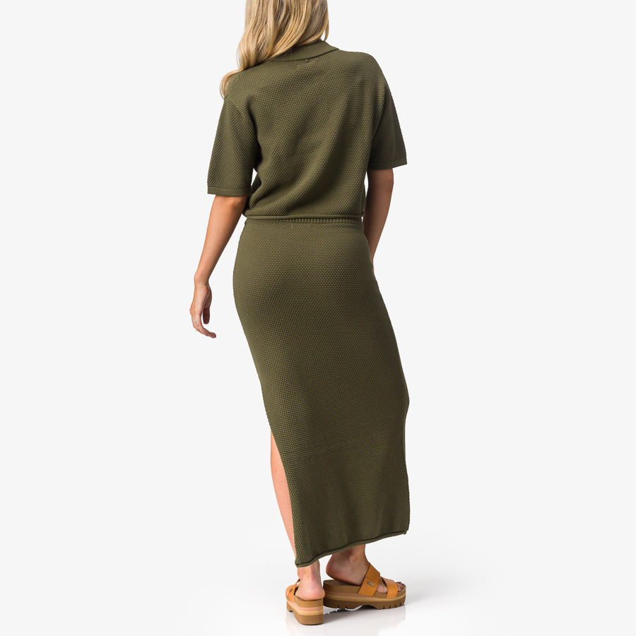 Woman wearing olive Up North Sweater Top shown from behind with soft fabric and tan sandals facing right for beach comfort