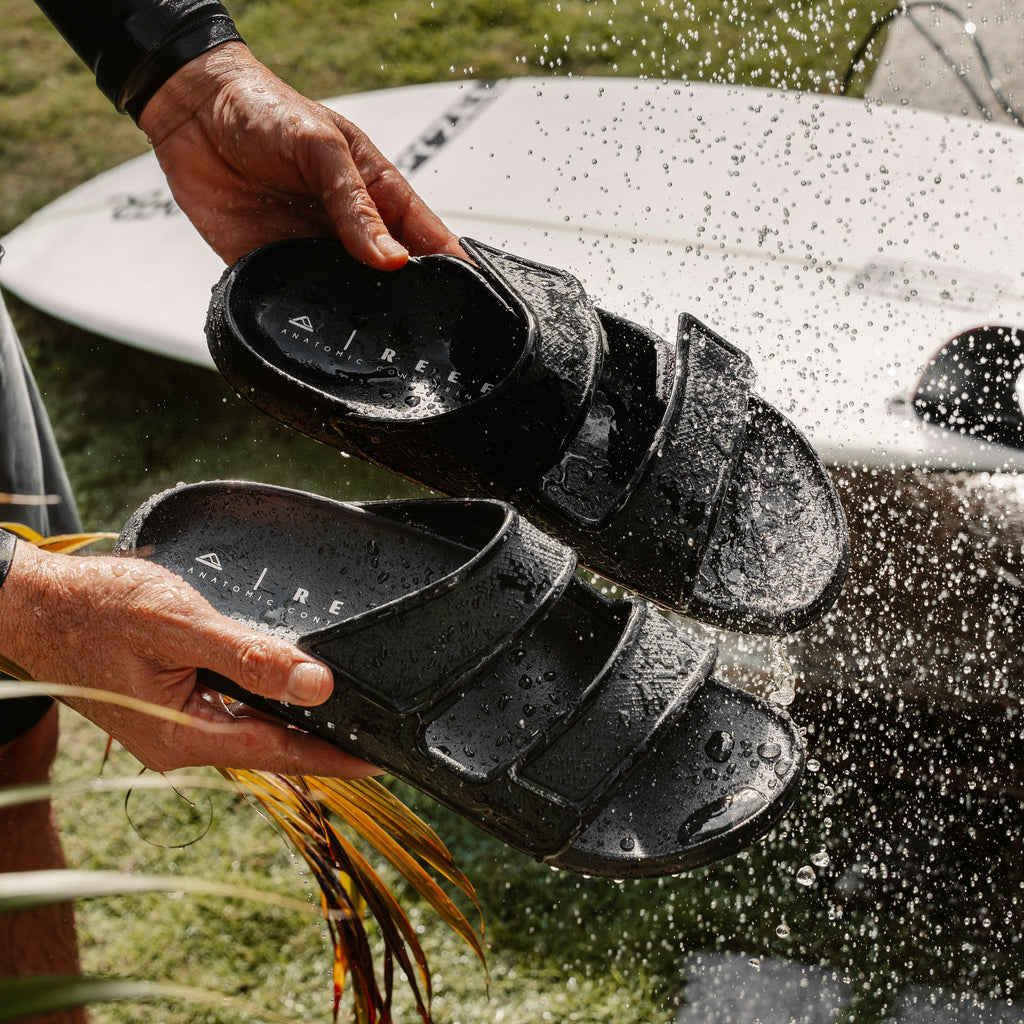 Oasis Adapt black men’s sandals facing upward in hands, water splashing, surf-inspired comfort with arch support