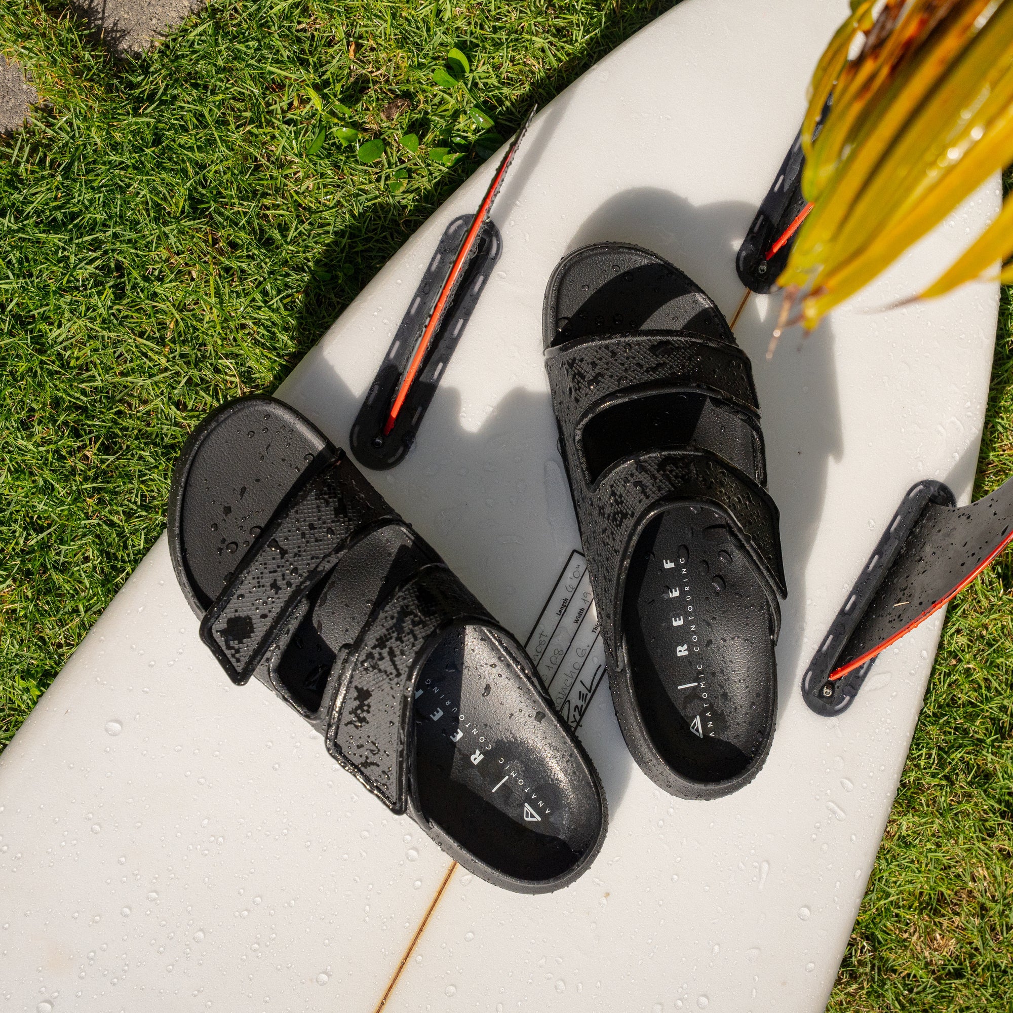 Oasis Adapt sandals facing up, wet water-friendly black shoes with adjustable straps on a white surfboard, beach-ready comfort.