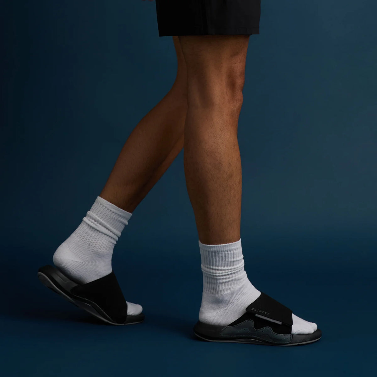 Cruiser Slide black men's footwear with anatomical arch support, facing left on a model wearing white socks against blue background.