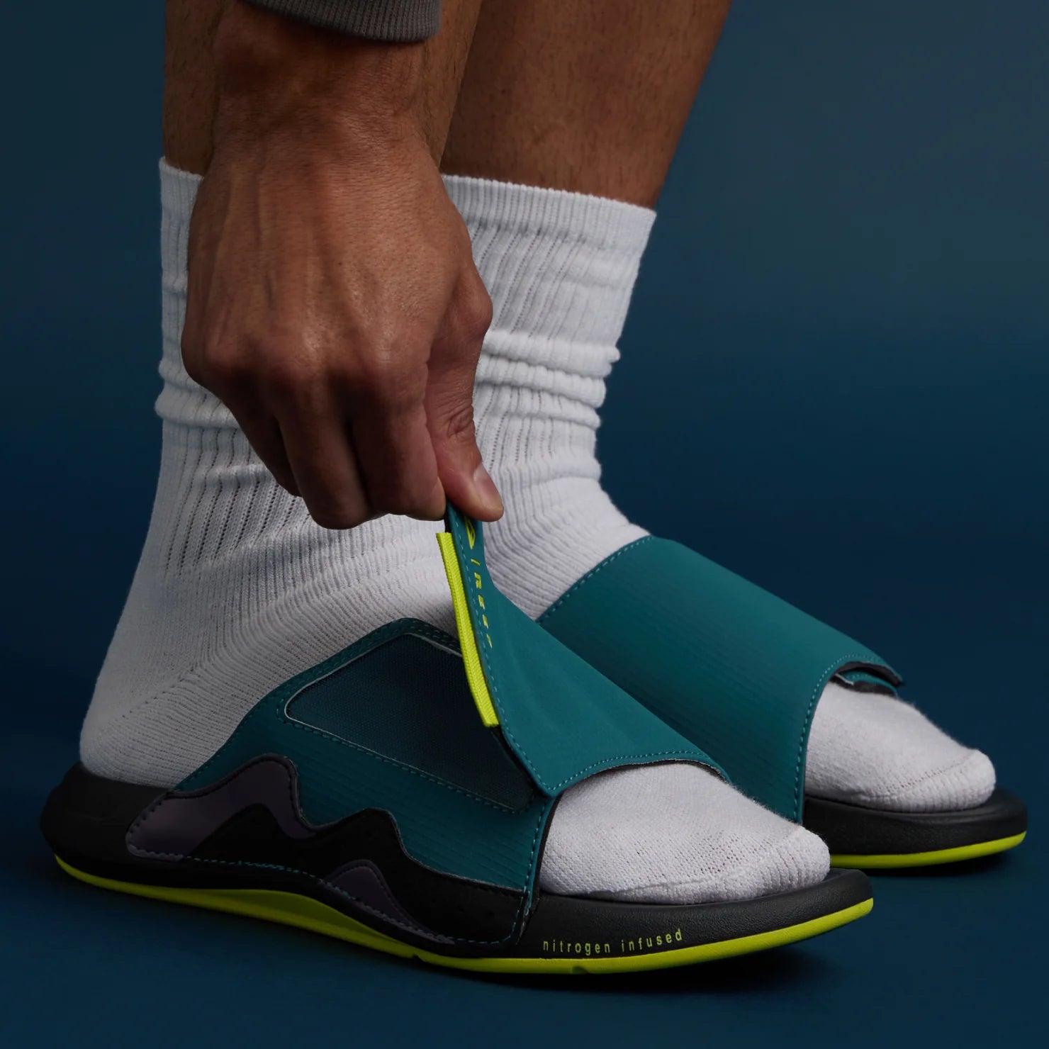 Man wearing white socks holding the front strap of a teal and lime Cruiser Slide facing forward, close-up on the foot and slide upper.
