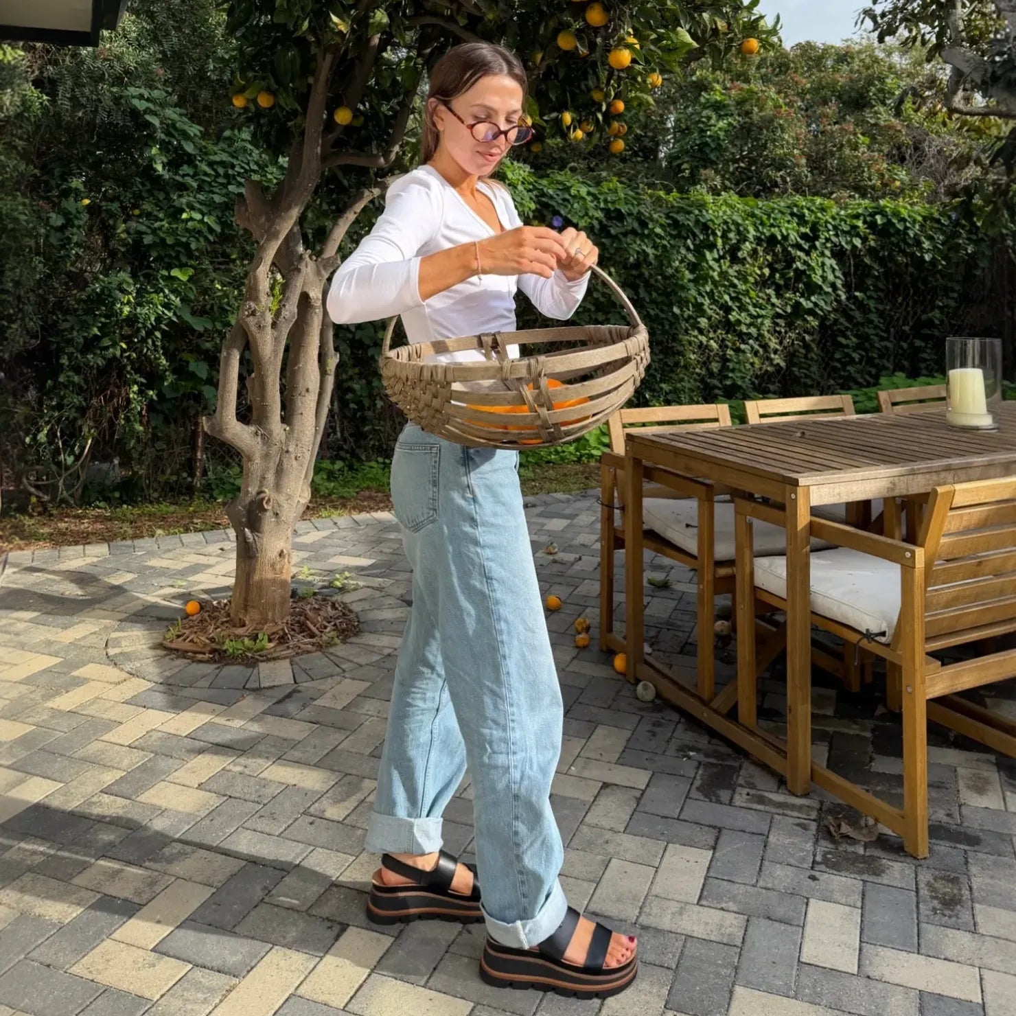 Woman wearing Vista Luxe Liah black sandals facing right, standing outdoors on stone patio near wooden table and orange tree