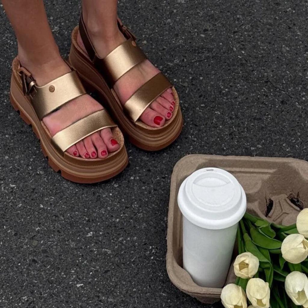 Vista Luxe Liah sandals facing forward, close-up of footbed and straps in warm bronze on asphalt with coffee and flowers nearby