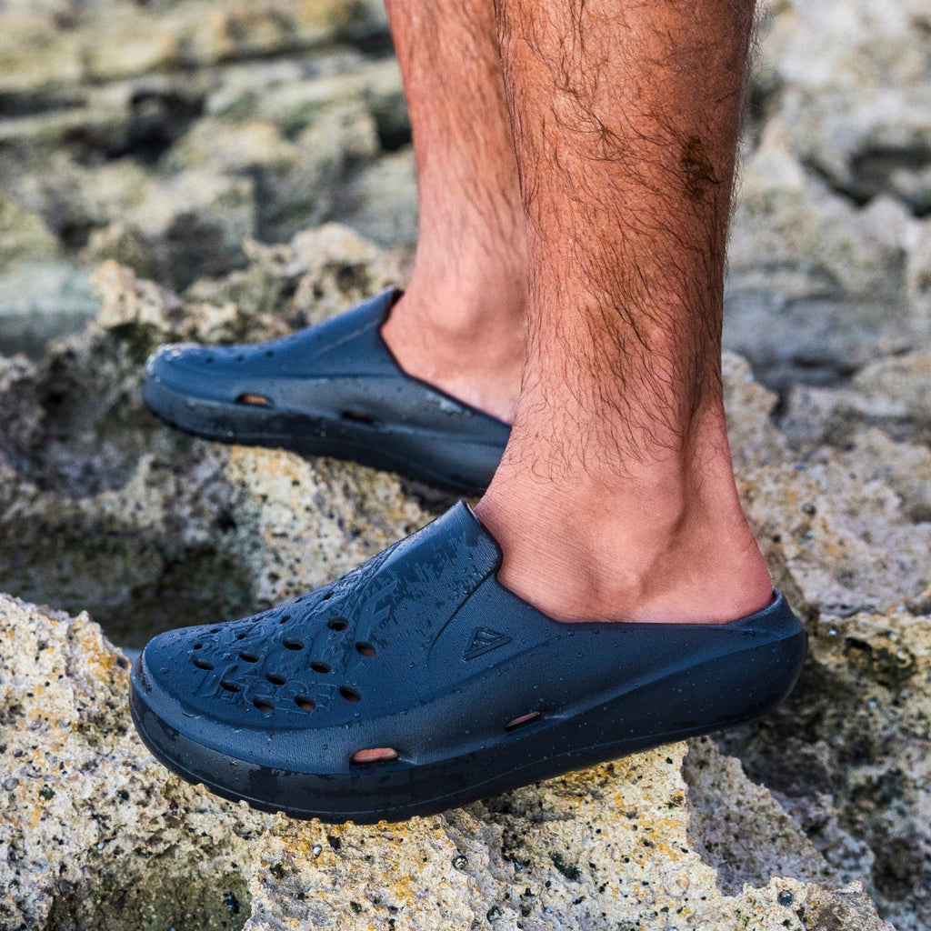 Glider men's footwear facing left, showing side view with water-friendly holes, soft cushioning on rocky beach surface.
