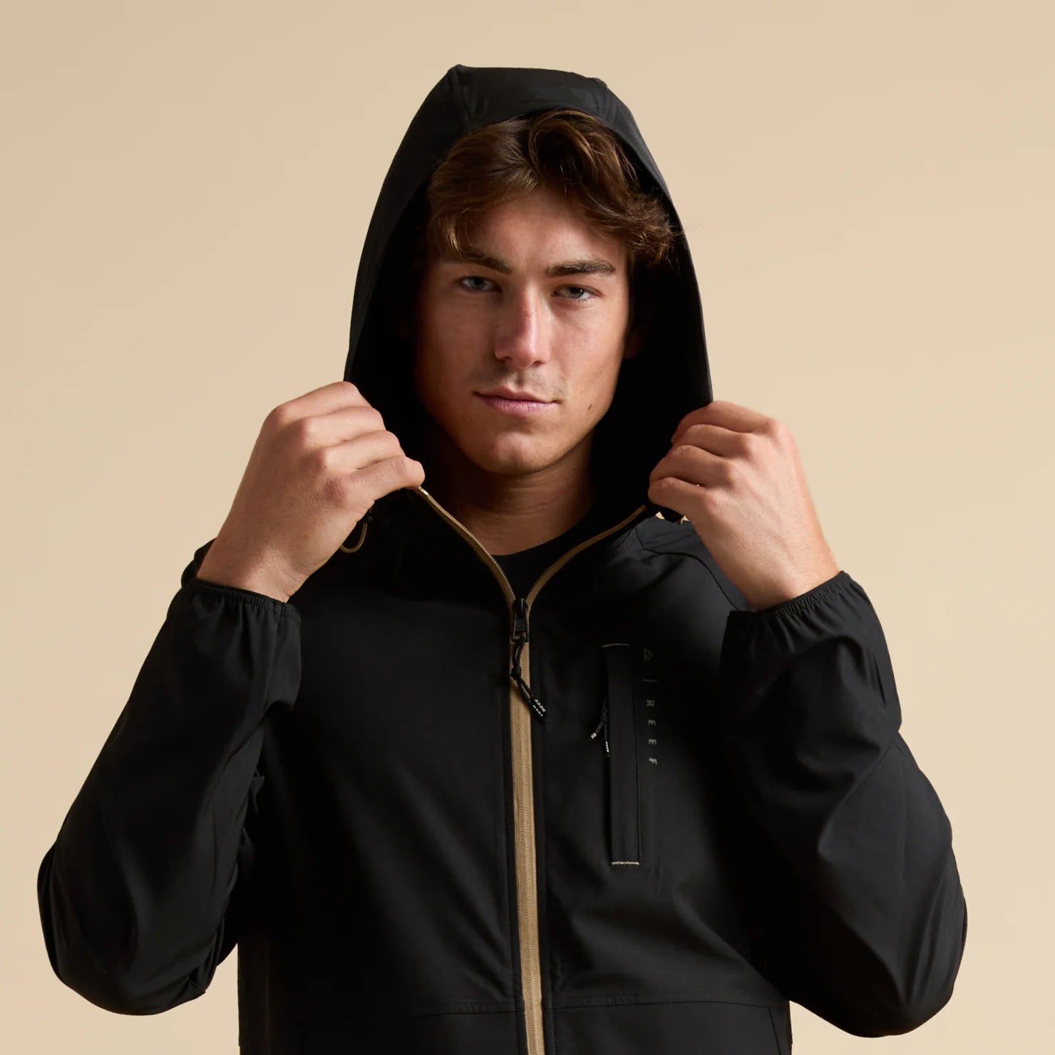 Man wearing a black Garvey Full Zip Jacket with hood up, front view showing front zipper and chest pocket details.