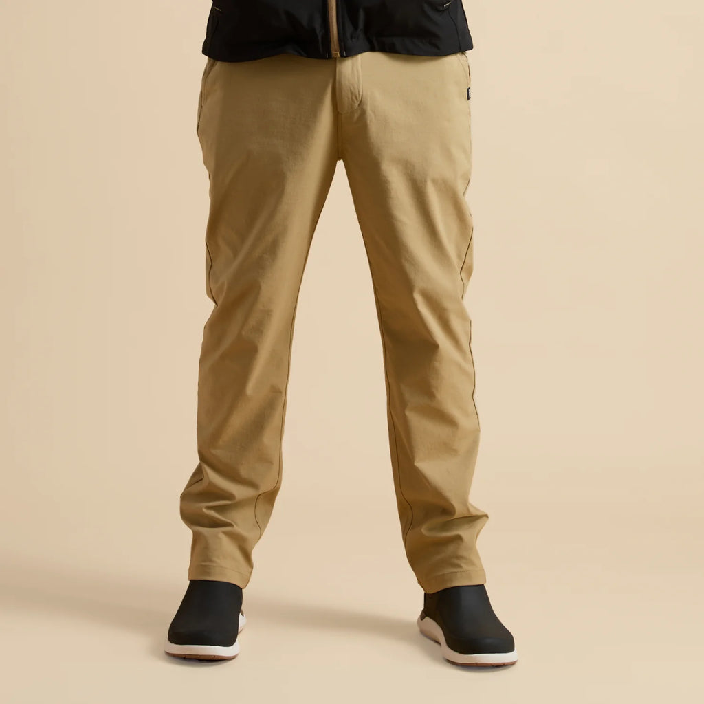 Front view of men's Carrick Stretch Pant in beige paired with black and white REEF slip-on shoes, beach-inspired comfort.