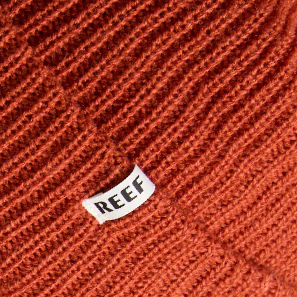 Close-up of textured chili-colored knit fabric with small white REEF tag on the unisex Alyx Beanie designed for beach comfort.