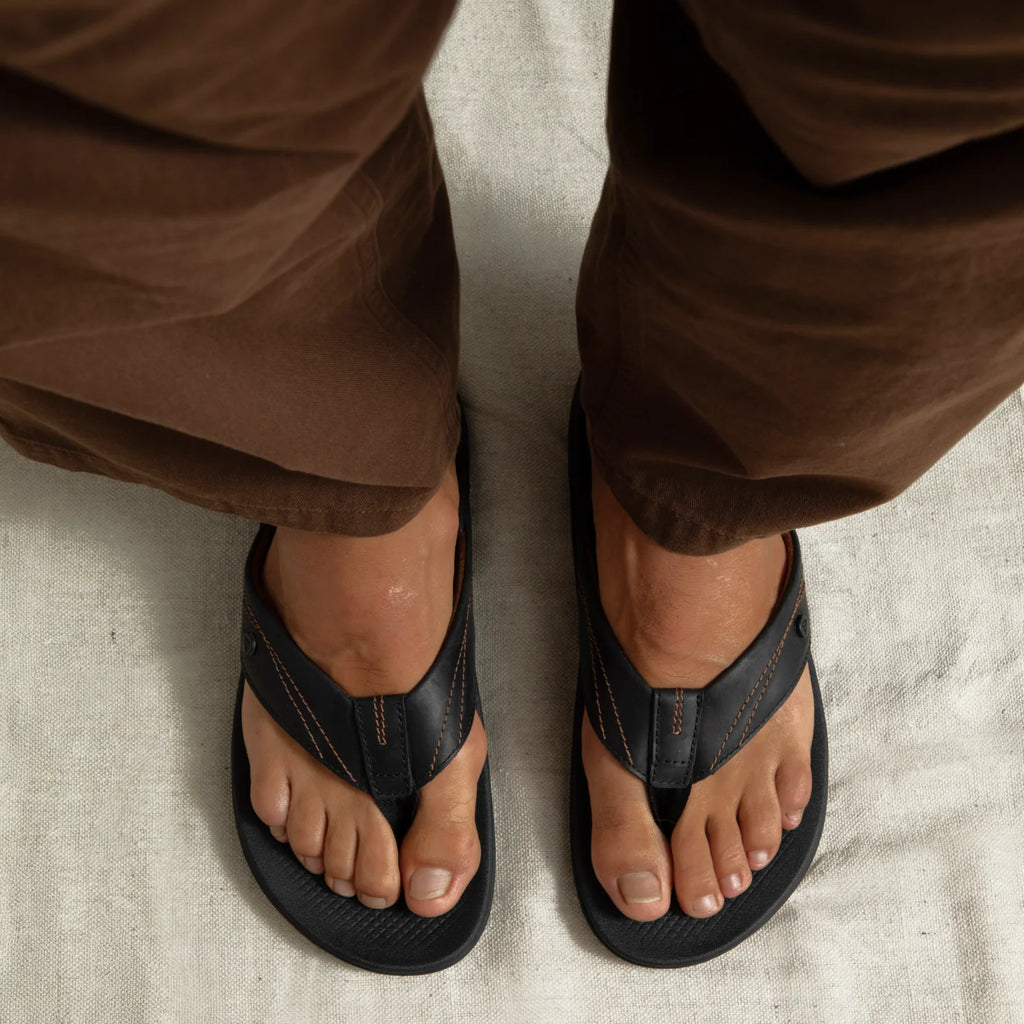 Top view of adult men's Phantom Lux black flip flops with anatomical arch support and soft cushioning, worn with brown pants.