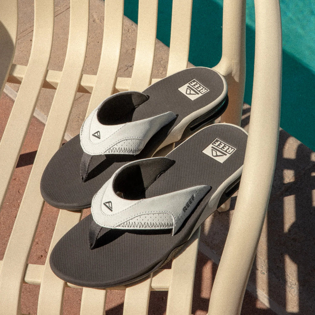 Pair of Fanning flip flops facing left with white synthetic nubuck straps, heel airbags, and black cushioned footbeds by REEF.