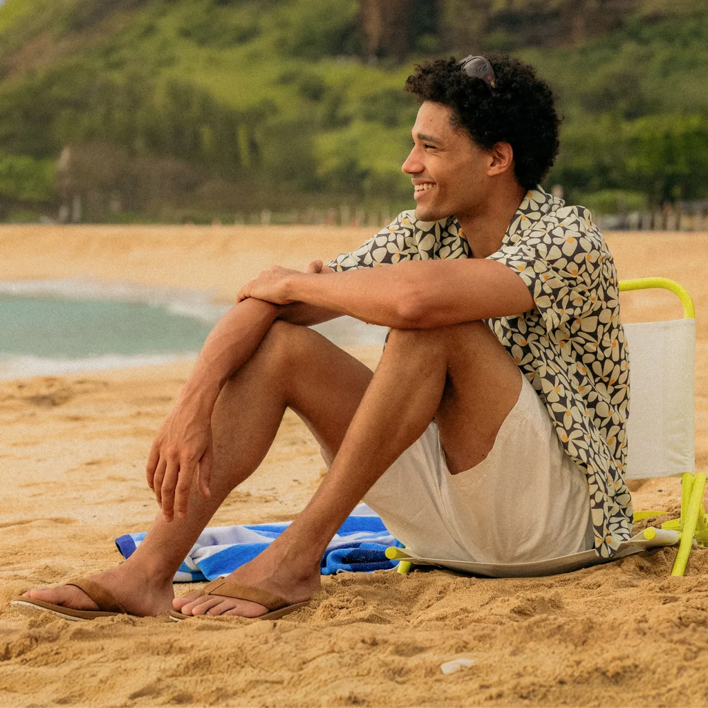 Man wearing Nox sandals facing left, sitting on sandy beach with surf-inspired comfort and arch support features.