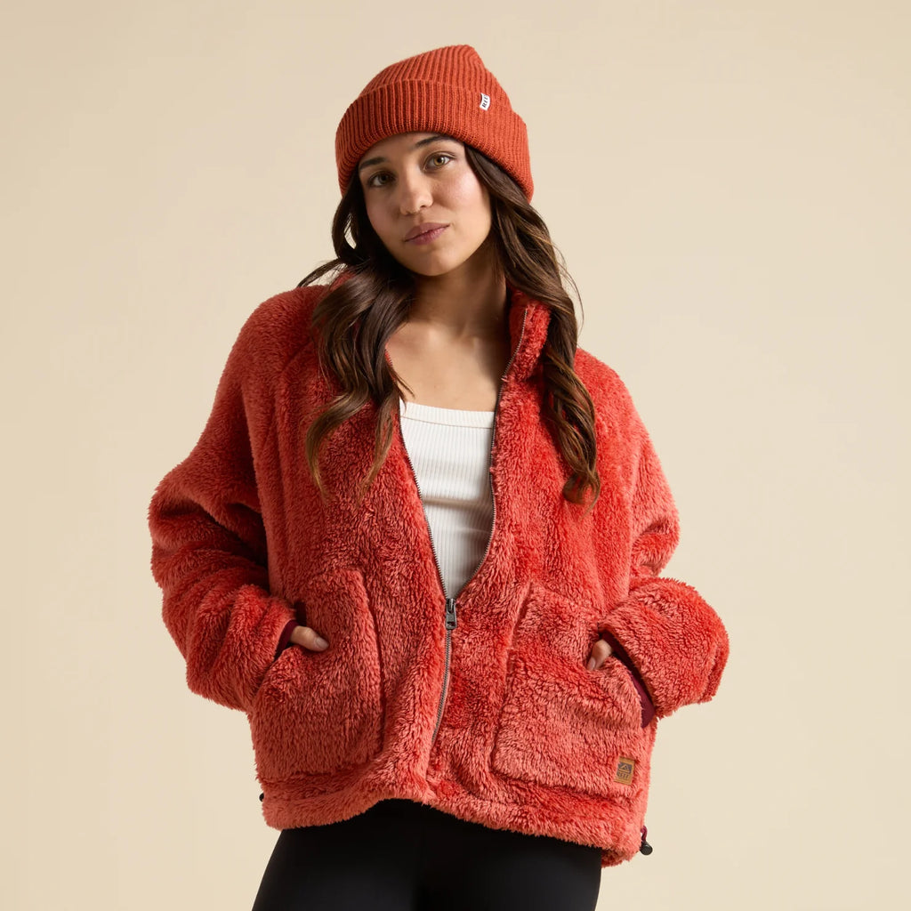 Woman wearing a coral Pilot Sherpa Jacket front view, hands in pockets, paired with a matching beanie, beach-inspired comfort apparel.