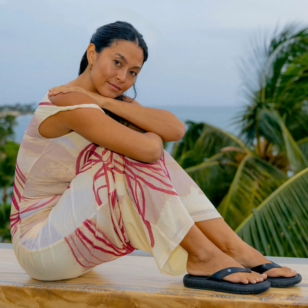 Woman sitting outdoors wearing Sunset Low Maya black sandals facing right, showing side and top views with a tropical background