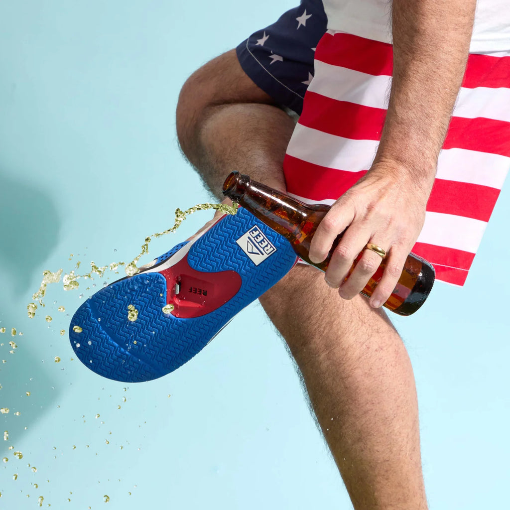 Close-up of the blue Fanning sandal sole facing upward with liquid splashing, worn by a man in patriotic shorts