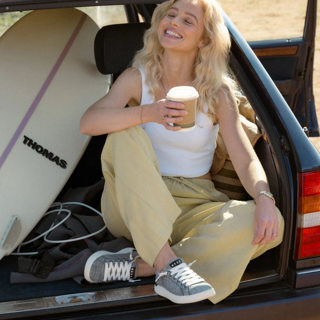 Woman wearing Neptune Alana sneakers facing forward, sitting in car trunk with surfboard, enjoying coffee and beach lifestyle.