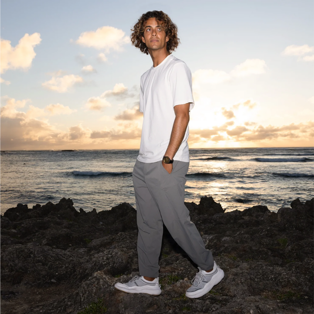 Man wearing Shorebreak TRV shoes facing left in grey walking pants, standing on rocky beach at sunset with ocean background.