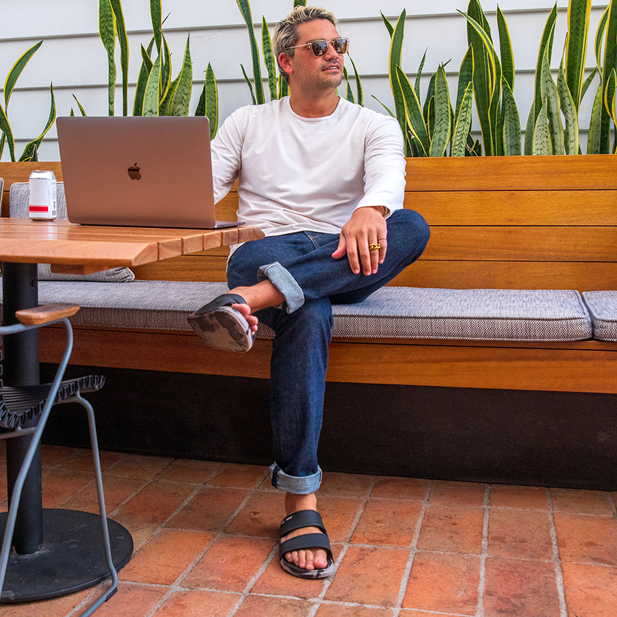 Oasis Double Up sandal on left foot facing forward, worn by man seated at outdoor table in casual setting