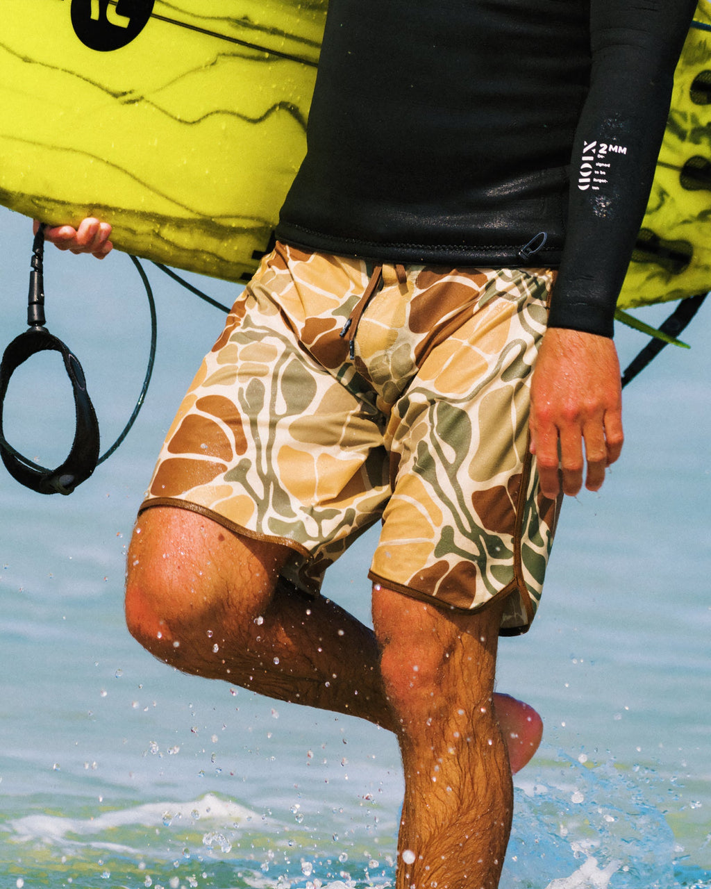 Man wearing Men's Palmera Boardshort - Floral Camo wading through water holding yellow surfboard, showcasing water-friendly fit and comfort.