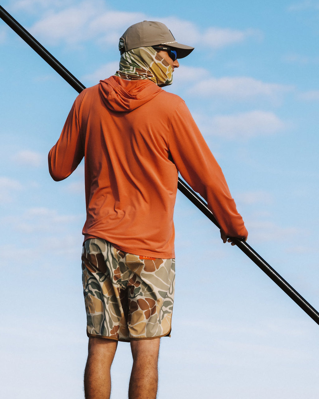 Man facing right wearing Men's Palmera Boardshort - Floral Camo with camo face covering and orange hoodie, holding black paddle outdoors