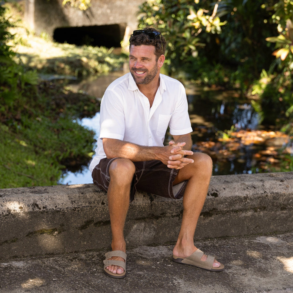 Man wearing tan Phantom Tradewind sandals facing forward, sitting outdoors near water, showcasing comfort and beach style.