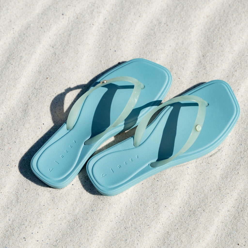 Solaye sandals facing right, close-up of blue sandals with translucent straps on sand, beach-inspired comfort design.
