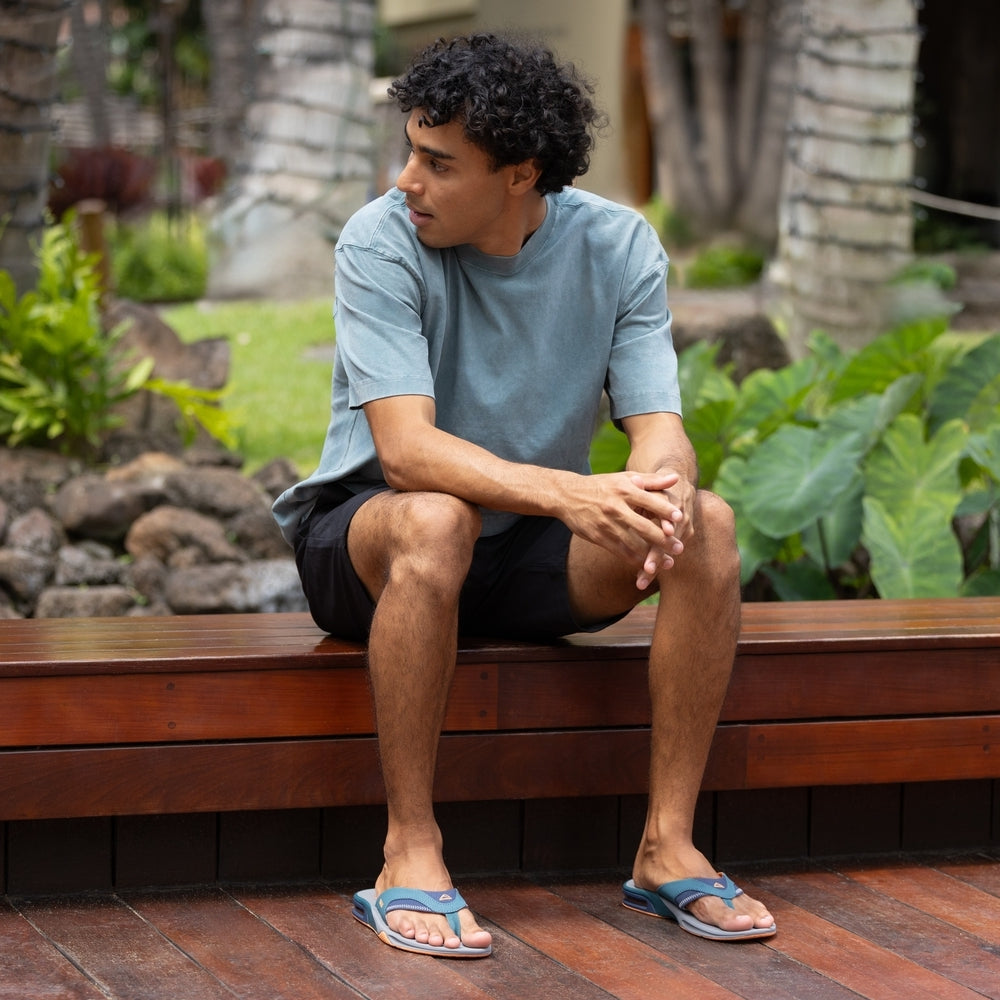 Man wearing Fanning sandals facing forward, seated outdoors on bench, showcasing comfort-focused beach-inspired footwear.