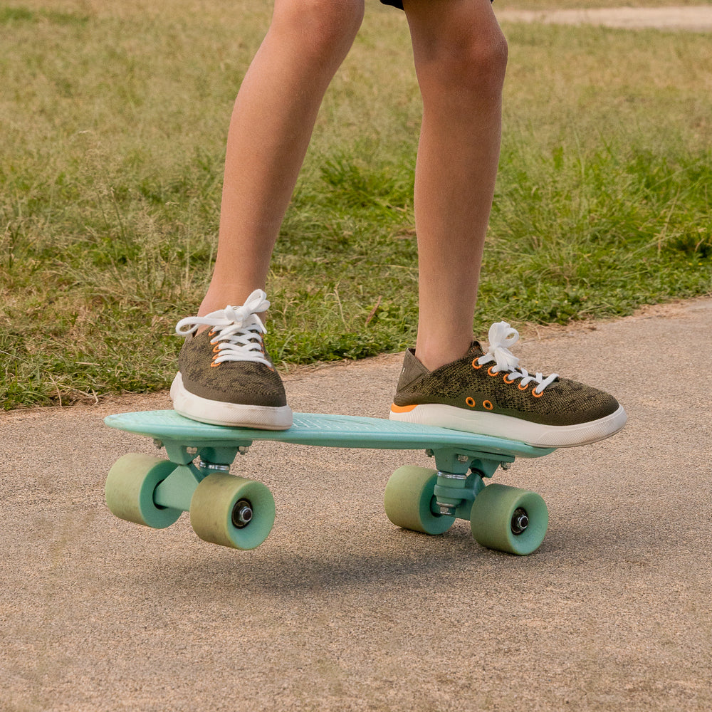 Kids Neptune shoes on a child’s feet facing forward, shown riding a teal skateboard outdoors on pavement.