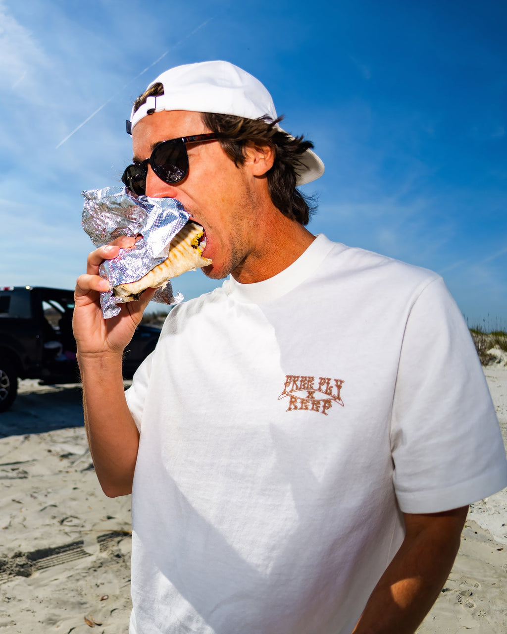 Man wearing High-Stoke Relaxed Tee - Bone facing left, eating, showing soft heavyweight cotton and laidback oversized fit on beach.
