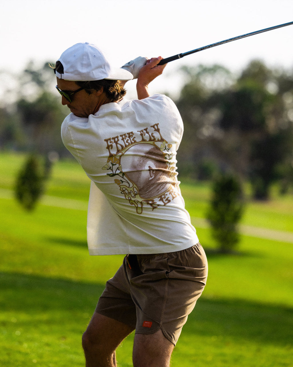 Man swinging golf club wearing High-Stoke Relaxed Tee - Bone with brown shorts, casual beach-inspired athletic look outdoors