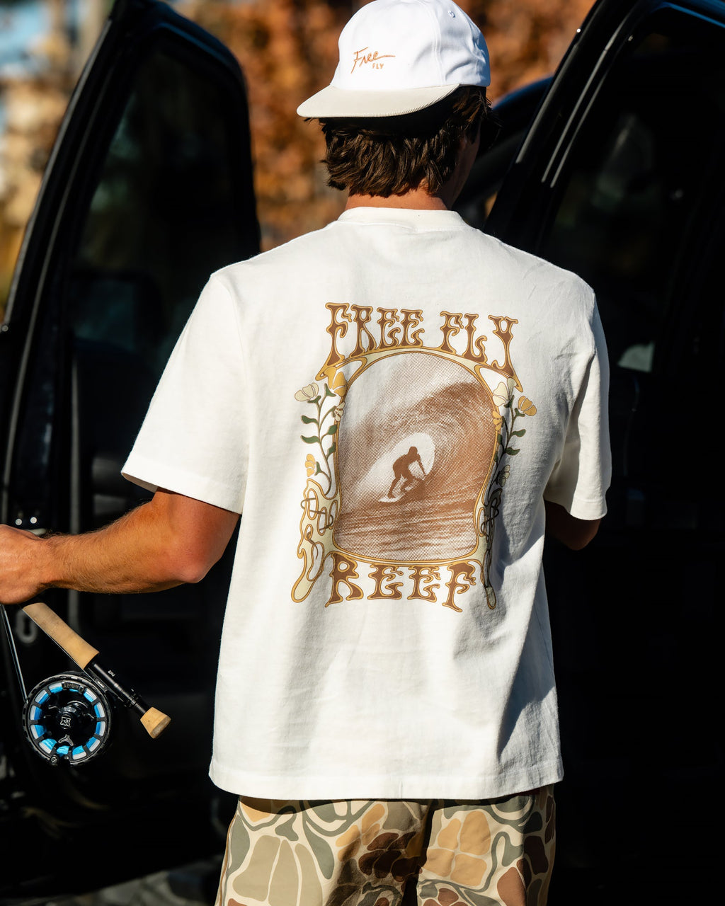 Man wearing High-Stoke Relaxed Tee - Bone showing back surf graphic, paired with camo shorts and white cap, beach lifestyle look
