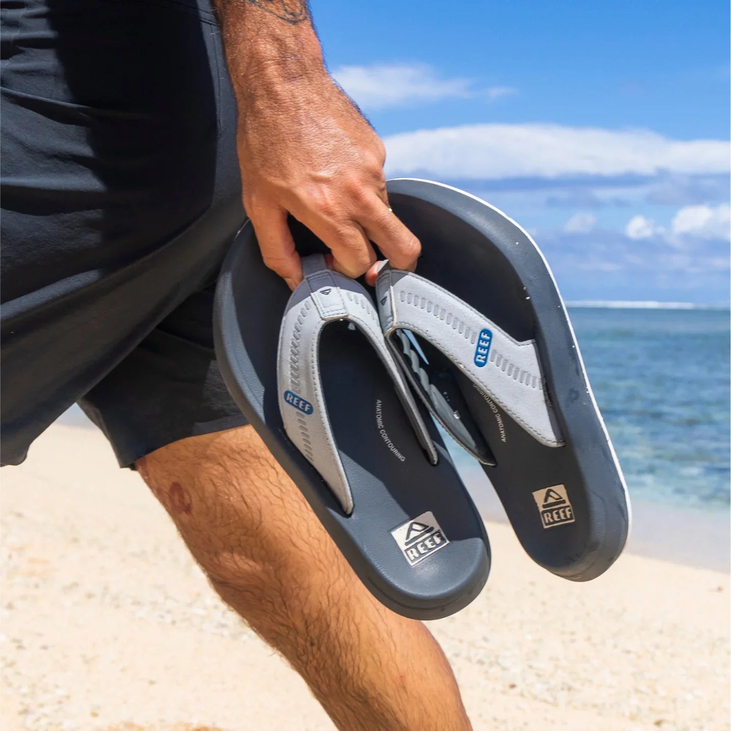 Man holding Cruiser flip flops facing downwards, showing the top straps with reef logos on a sandy beach background.
