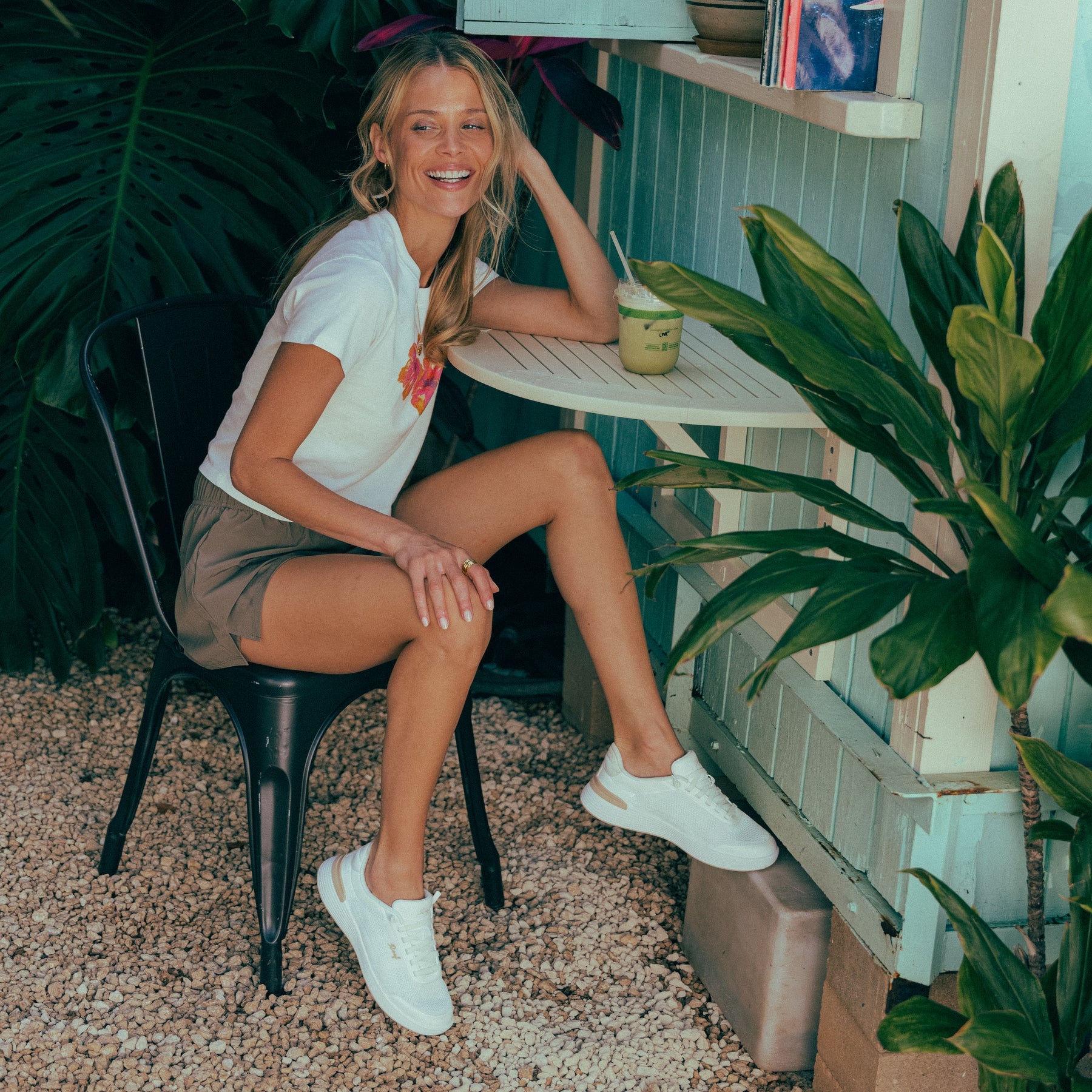 Woman wearing white Shoreline Palisades shoes facing right, seated outdoors with tropical plants and a drink on the table.
