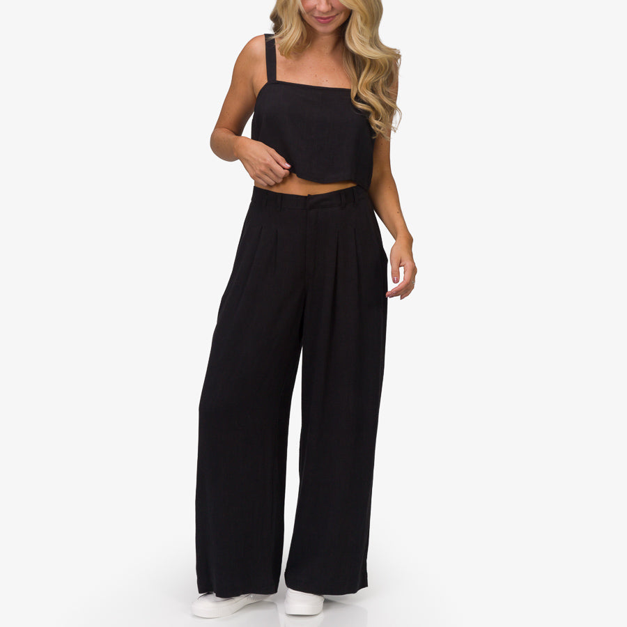 Woman wearing black Tori Linen Pant standing front view, showing full length and relaxed fit, paired with black crop top and white shoes.