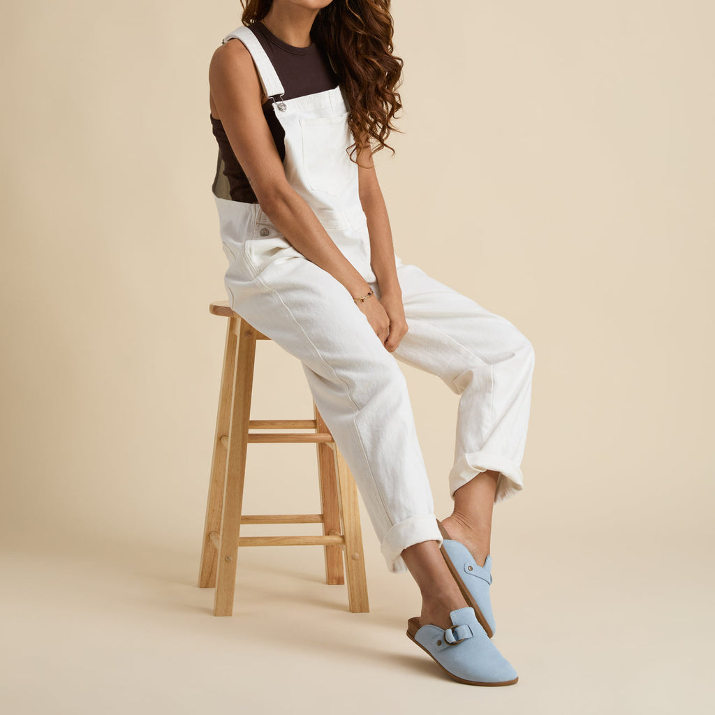 Woman seated on a stool wearing Vista Serenity dusty blue suede clogs facing left, highlighting soft cushioning and arch support.