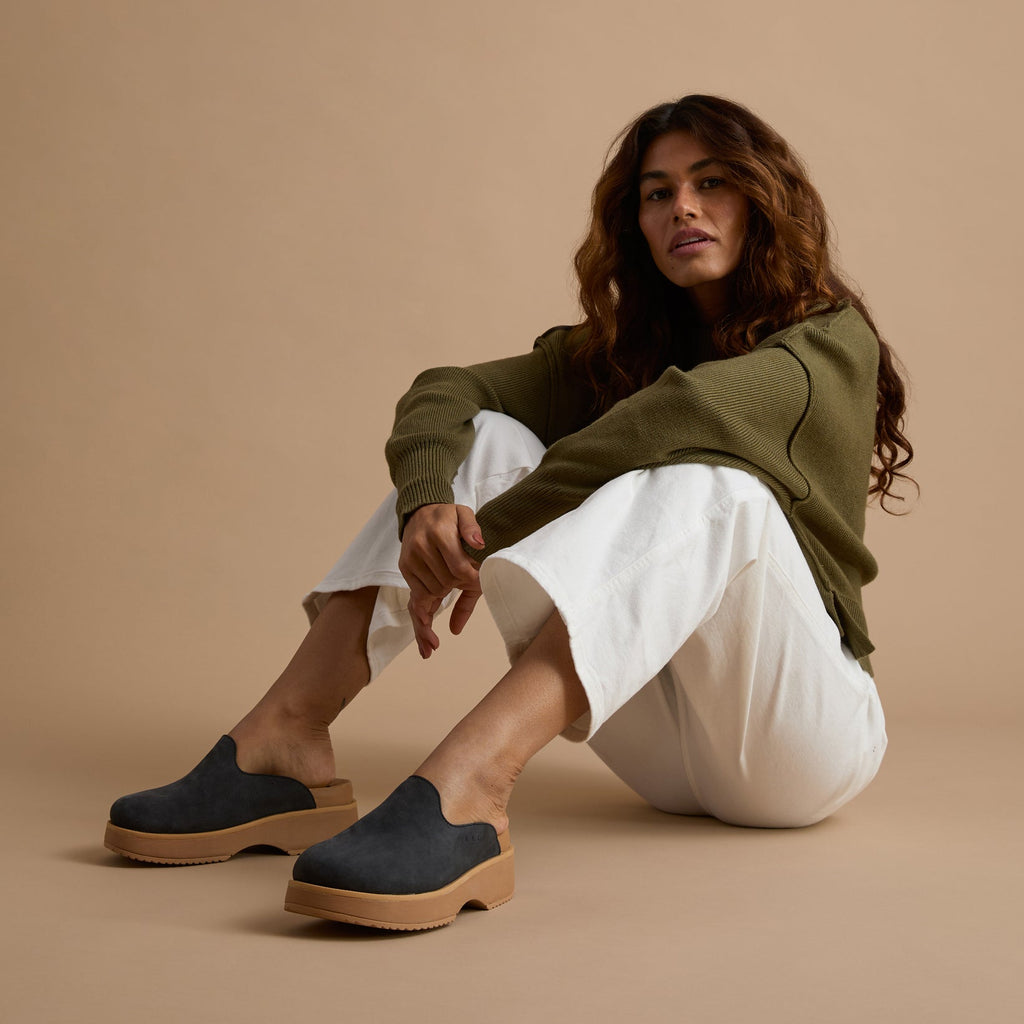 Vista Skye charcoal slip-on shoes facing left, shown from side on adult woman seated with white pants and green sweater